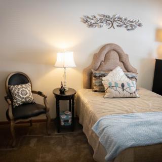A cozy bedroom with a classic design featuring a bed, nightstand with a lamp, and an elegant decorative chair.