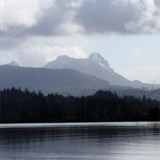 A serene view of a lake surrounded by forests and distant mountains under a cloudy sky.