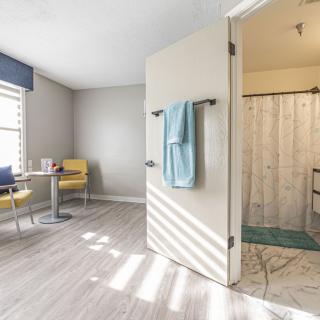 A modern interior space with a view into a bathroom, featuring light wood flooring, a small table with chairs, and a towel hanging on the door.