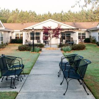 A serene outdoor scene with a pathway leading to houses, flanked by chairs and a well-maintained garden.