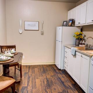 A small kitchen and dining area with wooden flooring, white cabinets, a refrigerator, and a table set for two.