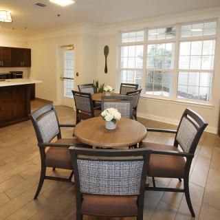 A cozy dining area with round tables and chairs, adjacent to a kitchen, featuring large windows that allow natural light to fill the space.
