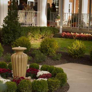 A meticulously landscaped garden with a central urn surrounded by vibrant flowers and neatly trimmed shrubs adjacent to a patio.