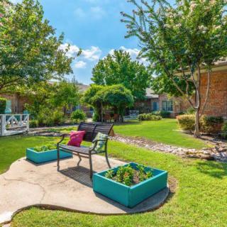 A peaceful garden with a bench surrounded by lush greenery and vibrant planters under a sunny sky.