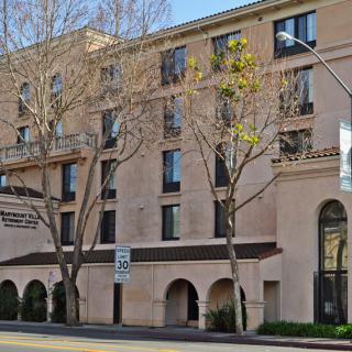 A city building with multiple floors, trees lining the sidewalk, and a clear blue sky above.