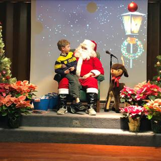 A child sits with Santa Claus on stage, surrounded by festive decorations and flowers.