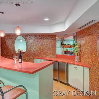 A stylish retro-themed kitchen bar area featuring orange tile walls, a pink countertop, modern pendant lights, and a bottle of wine on the counter.