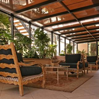 A cozy lounge area with wooden furniture and lush green plants under a glass ceiling.