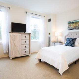 A bright and cozy bedroom with a white bed, dresser with a TV, and large windows accentuated by light curtains.