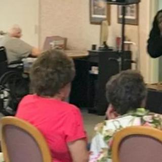 A group of elderly individuals gathers indoors, with one person in a wheelchair, likely in a nursing home setting.