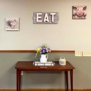 A cozy kitchen setup featuring a wooden table with a ceramic pitcher of flowers, surrounded by playful pig-themed wall art and a rustic 'EAT' sign.