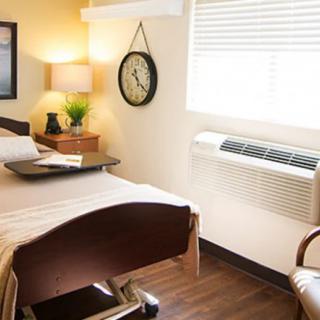 A clean and organized hospital room with a bed, side table, lamp, clock, and window.