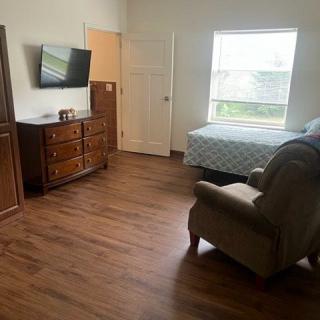A cozy bedroom with wooden flooring, featuring a bed, an armchair, a dresser, and a wall-mounted TV.