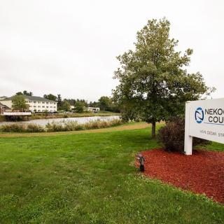 A scenic view of a park featuring a sign, a tree, grassy area, and a lake with buildings in the background.