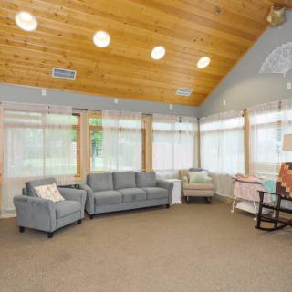 A cozy living room with a wooden ceiling, featuring a sofa, armchair, and large windows.