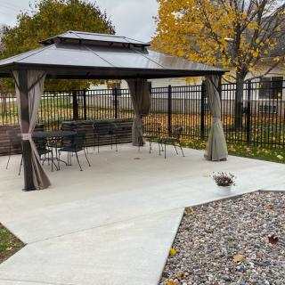 A gazebo with outdoor furniture on a patio, surrounded by autumn trees and a fence.