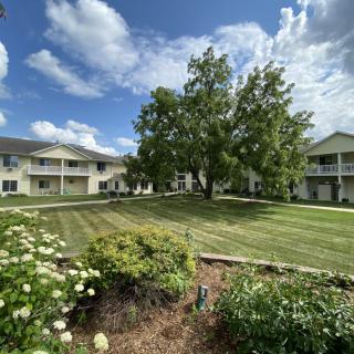 A serene view of an apartment complex with well-maintained gardens and a large tree under a partly cloudy sky.