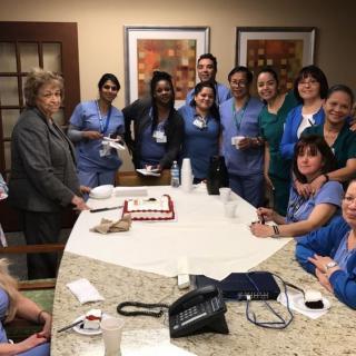 A group of healthcare professionals in scrubs gathered around a table in an office setting, possibly celebrating an occasion.