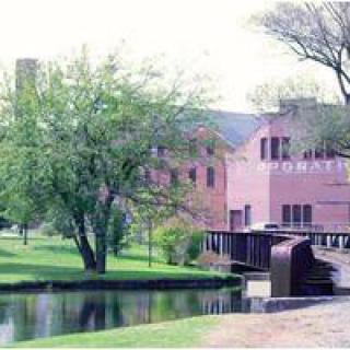 A serene landscape featuring a brick building, a small bridge over water, and lush green trees.