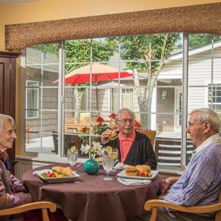 Three seniors enjoying a meal together by a large window overlooking a patio.