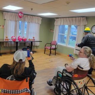 A group of people in wheelchairs participating in an indoor activity, with balloons and decorations visible in the background.