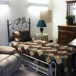 A cozy bedroom featuring a quilt-covered bed, a lamp on a side table, a comfortable chair, and a wooden dresser.