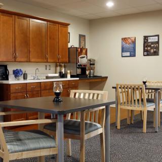 A cozy breakroom with wooden cabinets, tables and chairs, equipped with a coffee machine and decorated with wall posters.