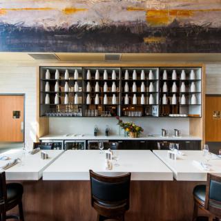 A modern restaurant interior featuring a bar with seating and neatly arranged shelves of glasses.