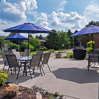 A sunny patio with blue umbrellas and outdoor seating, surrounded by a landscaped garden.