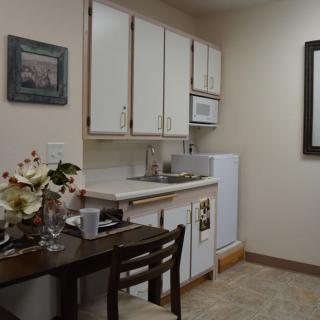 A small kitchen area with cabinets, a sink, microwave, refrigerator, and a table with chairs and decorative flowers.