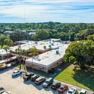 An aerial view of a building surrounded by trees with a parking lot filled with vehicles on a sunny day.