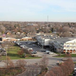 A suburban area with buildings and a parking lot, surrounded by trees and rooftops under a clear sky.