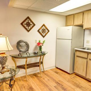 A cozy kitchen area with wooden floors, a fridge, and decorative elements such as a table with flowers, a lamp, and wall art.