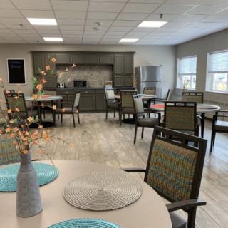 A modern dining area with neatly arranged tables and chairs, featuring a view of the kitchen in the background.