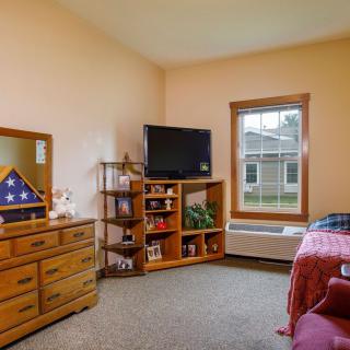 A cozy bedroom with a neatly arranged dresser, television, and a window overlooking the outside.