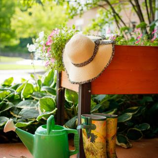 A sun hat and a green watering can next to a raised planter filled with blooming flowers.
