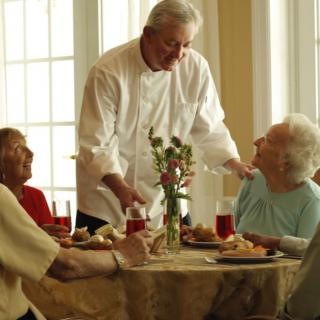 An elderly group enjoys a meal served by a waiter in a bright dining setting.
