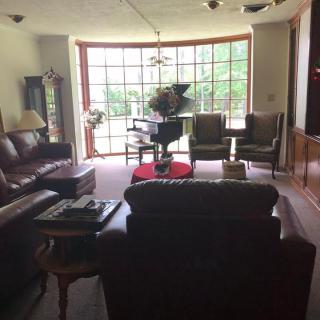A cozy living room with a grand piano by a large window, surrounded by comfortable seating and tasteful decor.