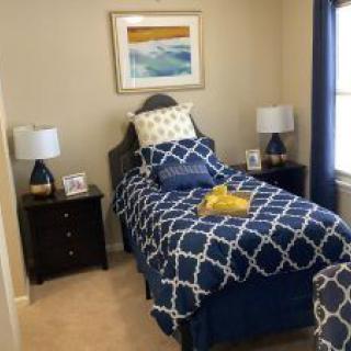 A neatly arranged bedroom with a single bed featuring blue patterned bedding, flanked by bedside tables and lamps, and a piece of artwork hanging above.