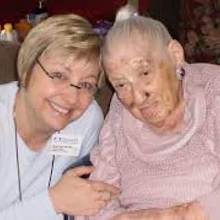 A caregiver smiling and posing with an elderly woman indoors, showcasing companionship.