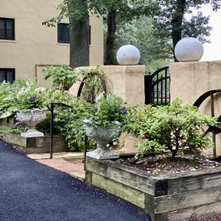 A charming garden with lush greenery, decorative urns, and architectural elements along a paved pathway.
