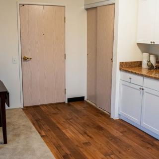A small kitchen area with wooden flooring, white cabinets, and a countertop adjacent to a doorway.