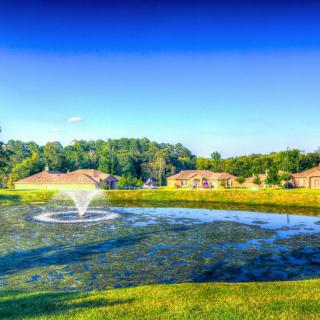 A picturesque view of a pond with a fountain surrounded by houses and lush greenery under a clear blue sky.