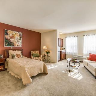 A cozy bedroom with a bed, chairs, and a small kitchenette, bathed in natural light.