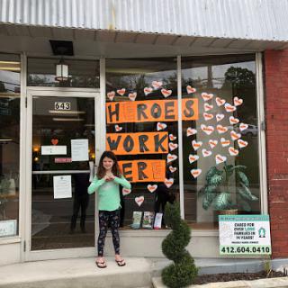 A child stands in front of a store with a colorful window display celebrating heroes and community workers.