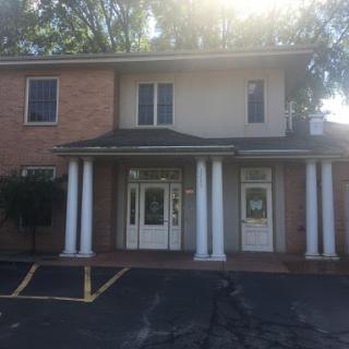 A two-story brick building featuring a welcoming entrance with columns, illuminated by sunlight.