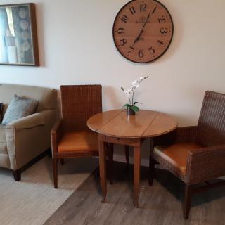 A cozy living room corner featuring two chairs, a round wooden table with a decorative flower arrangement, and a wall clock above.