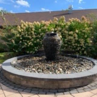 A garden fountain with an urn centerpiece surrounded by plants and a paved patio.