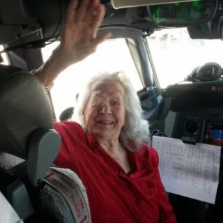 Elderly woman smiling and waving while seated in an airplane cockpit.