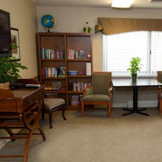 A cozy reading area featuring a bookshelf, comfortable chairs, and a small table, illuminated by natural light.